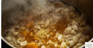 Chopped onions, garlic, and ginger sautéing in a large pot with oil. The aromatics are softening and releasing fragrant steam, creating a flavorful base for Jamaican Curry Chicken.