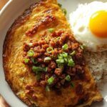 Golden-brown roasted and flattened tortang talong with giniling - eggplant omelet topped with cooked ground pork, garnished with green onions, served with garlic rice and a sunny-side-up egg on a white plate, Filipino breakfast.
