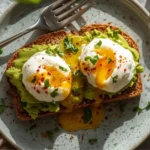 Creamy avocado toast with poached egg on whole-grain bread, garnished with microgreens and red pepper flakes.